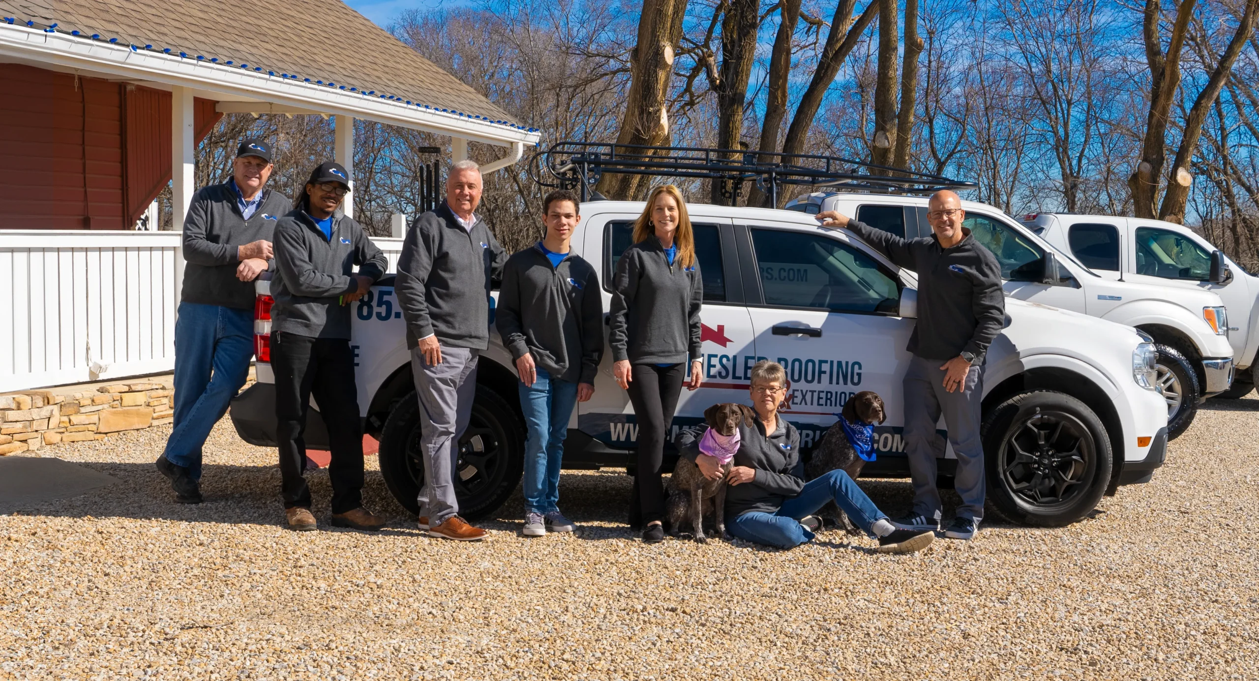 Mesler Roofing & Exteriors team standing in front of branded service trucks with two dogs outside a home